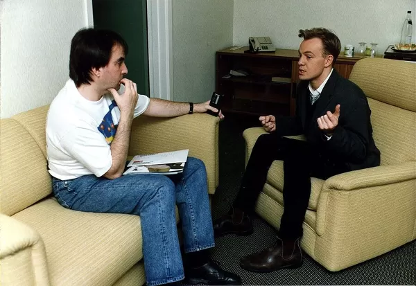 Jason Donovan being interviewed black jeans jacket sitting in armchair ankle boots